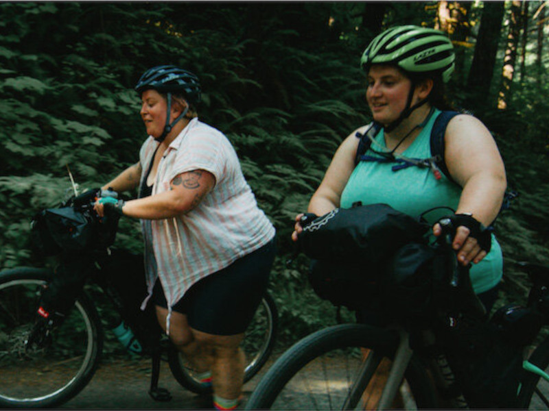 All Bodies on Bikes brand image: Two plus sized women ride bikes through the forest.