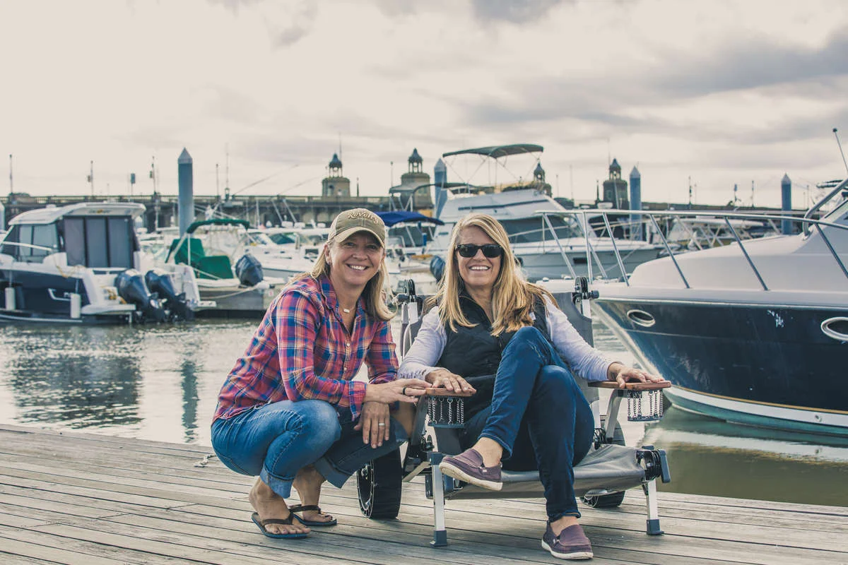 Do It In Nature Podcast brand image: Two women sit on a dock in a marina with big grins on their faces.
