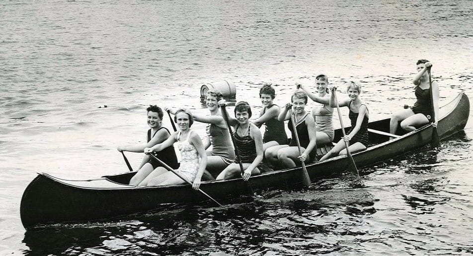 Camp Newaygo brand image: A vintage photo of several women paddling and smiling together in a canoe.