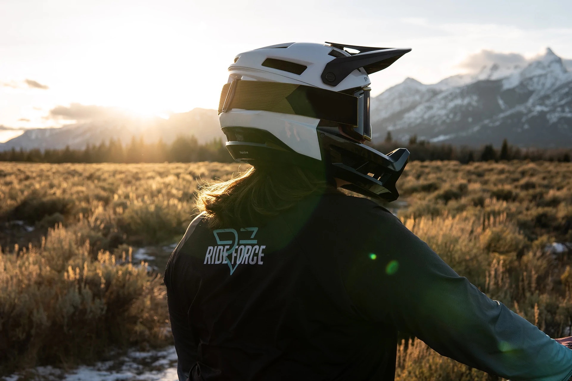 A mountain biker wears a helmet and Ride Force jersey looks out over a mountain valley at sunset.
