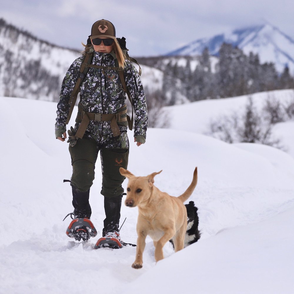 Woman walking with a dog through snow, wearing camouflage hunting apparel.