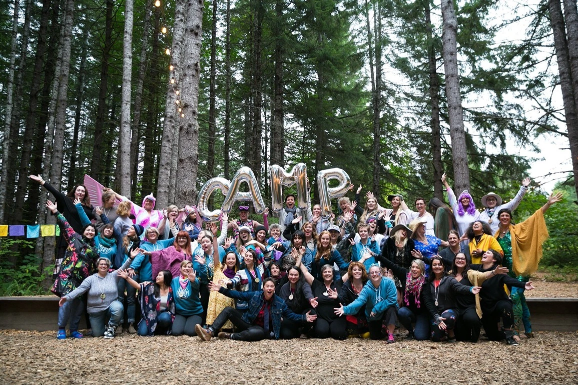 a large group of women gather together in a forest holding balloons that spell "camp"