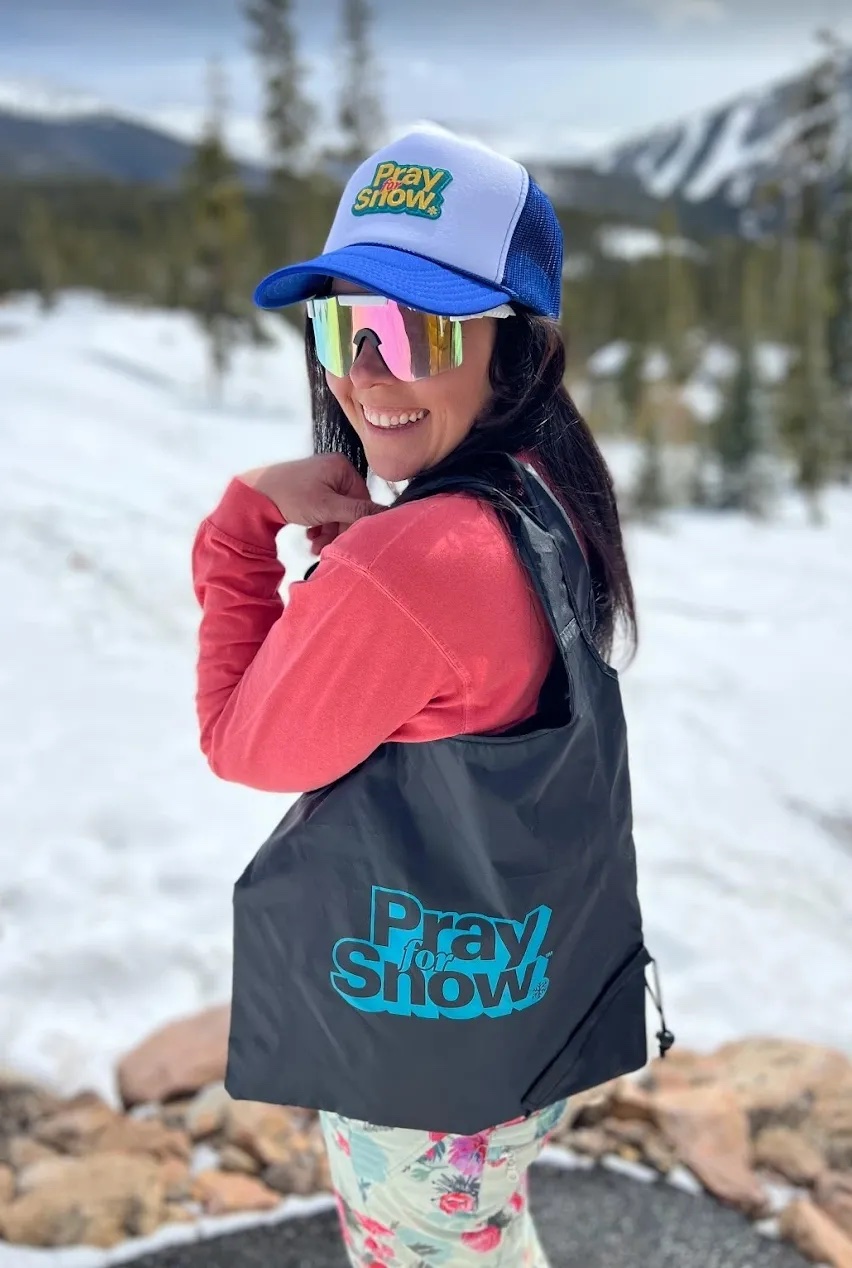 a woman in sunglasses and hat shows off a tote bag with the words "pray for snow" on it