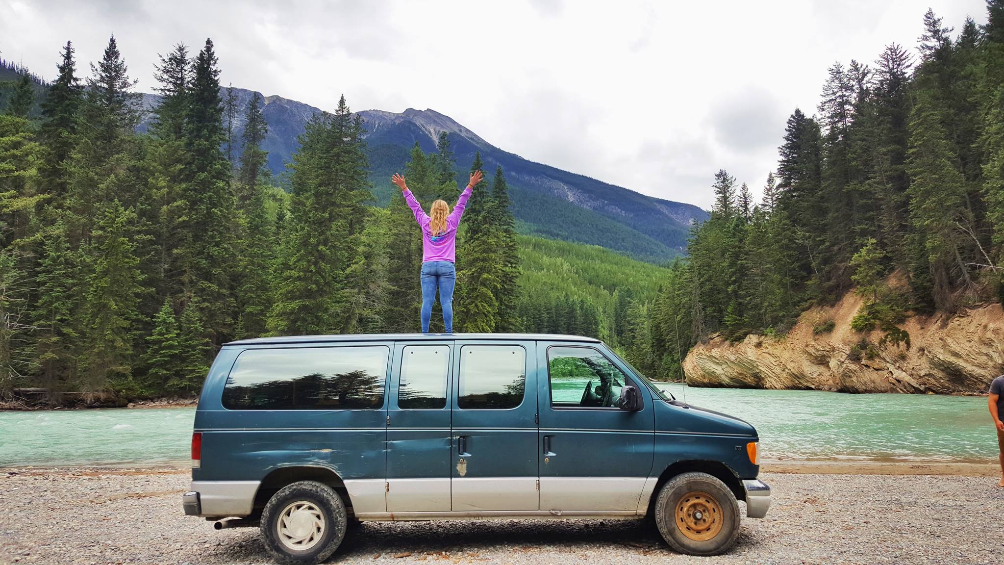 a woman stands on top of a van in a pretty nature setting